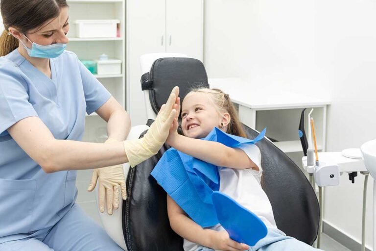 Parent helps a young child brush teeth at a bathroom sink with a soft toothbrush and fluoride toothpaste.